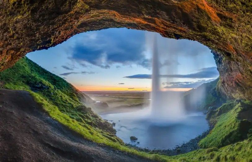 Waterfall viewed from within a cave; water cascades into a pool. Green vegetation frames the opening, cloudy sky visible.