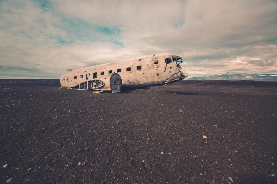 Wreckage of a white airplane on a black sand beach under a cloudy sky.
