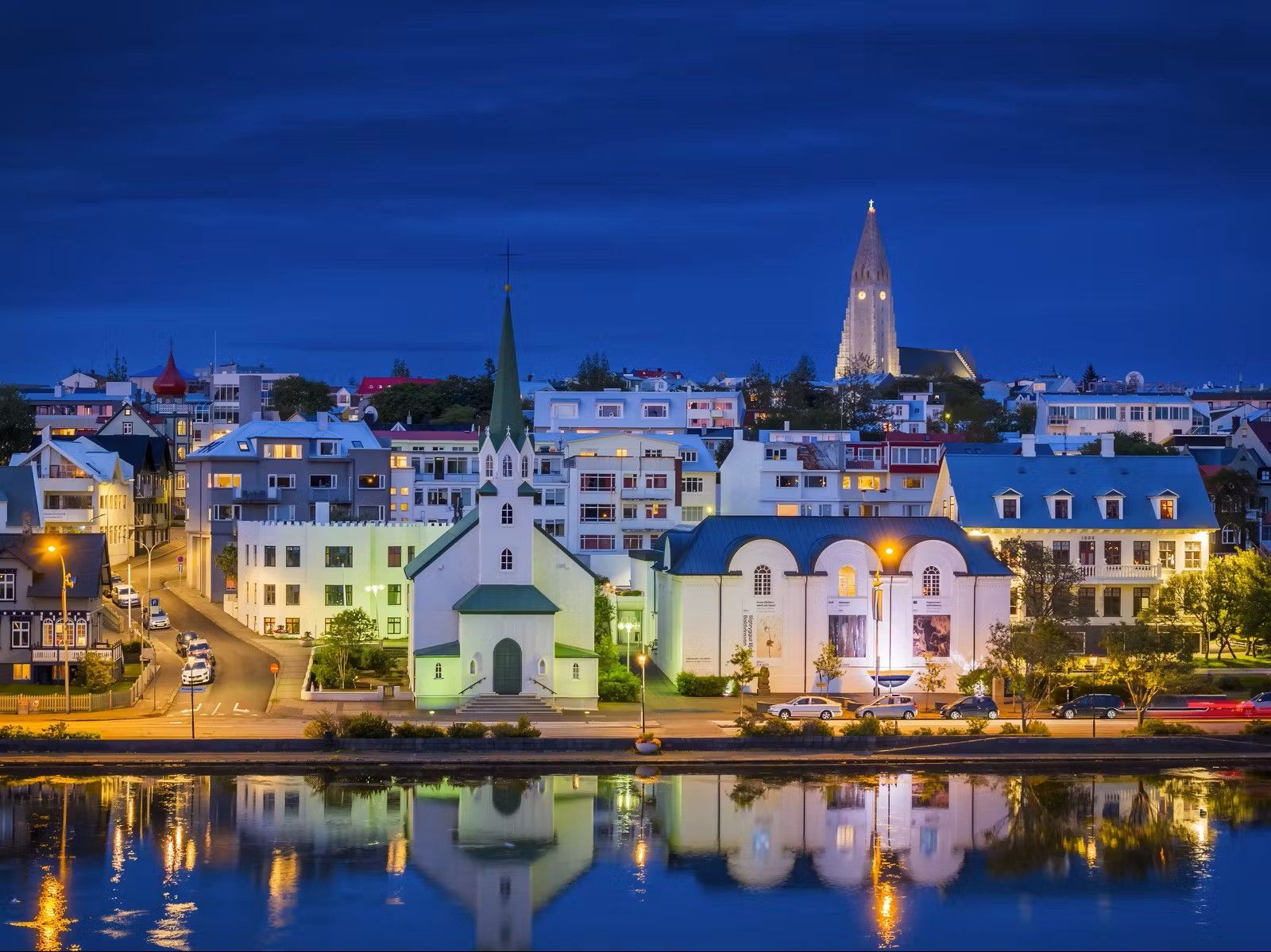 Cityscape at dusk, illuminated buildings reflected in water; church spires rise against a dark blue sky.