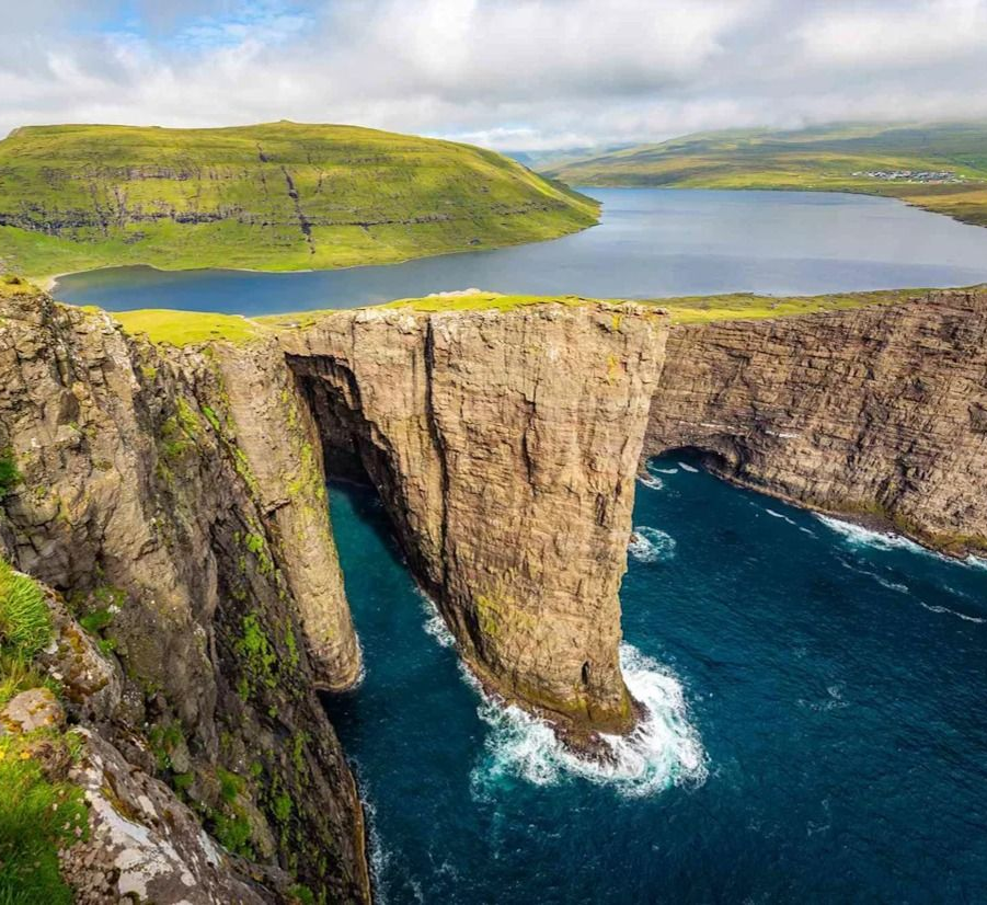 Cliffs overlooking blue ocean and a lake, lush green hills in the background under cloudy skies. The one of its kind Lake Sorvagsvatn