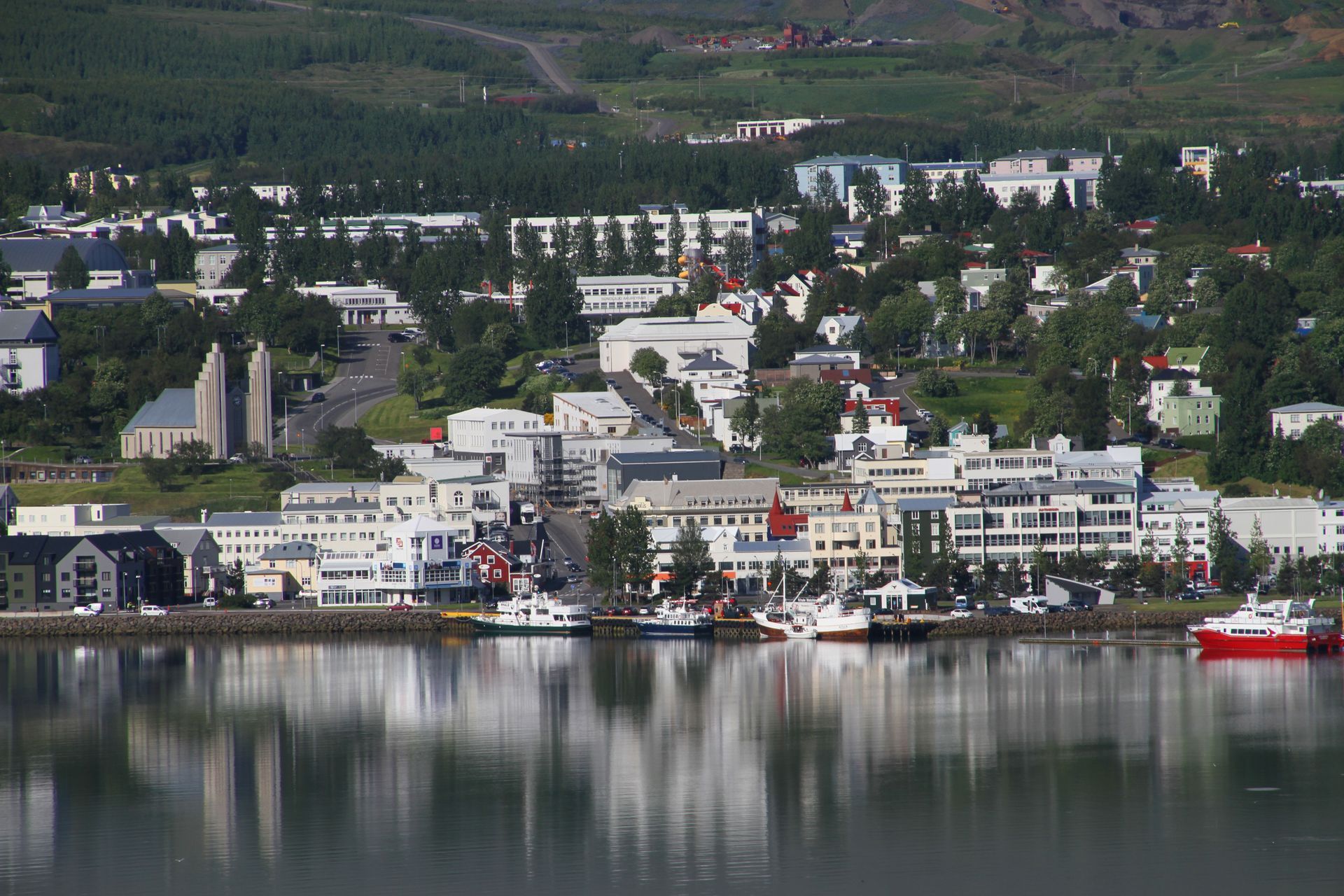 Town waterfront with buildings and boats reflecting in the water, under a green hillside.