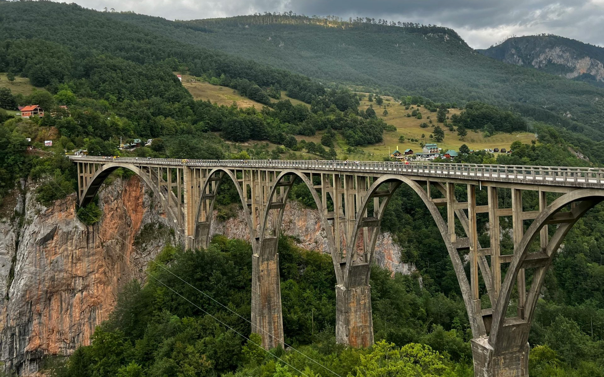 Concrete arch bridge spans a deep canyon, surrounded by green mountains and a cloudy sky.