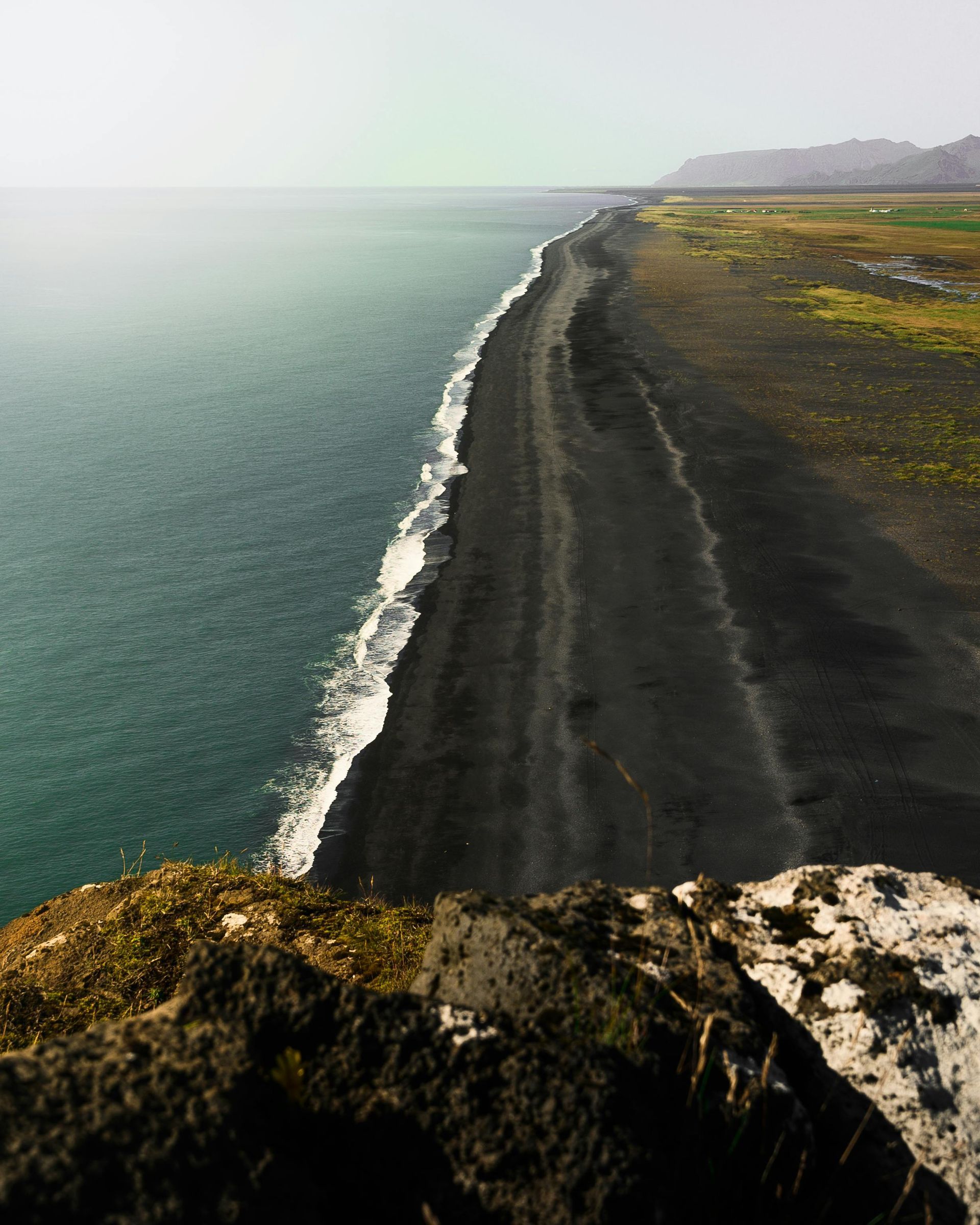 Black sand beach with teal ocean, contrasting with a green-brown landscape under a hazy sky.