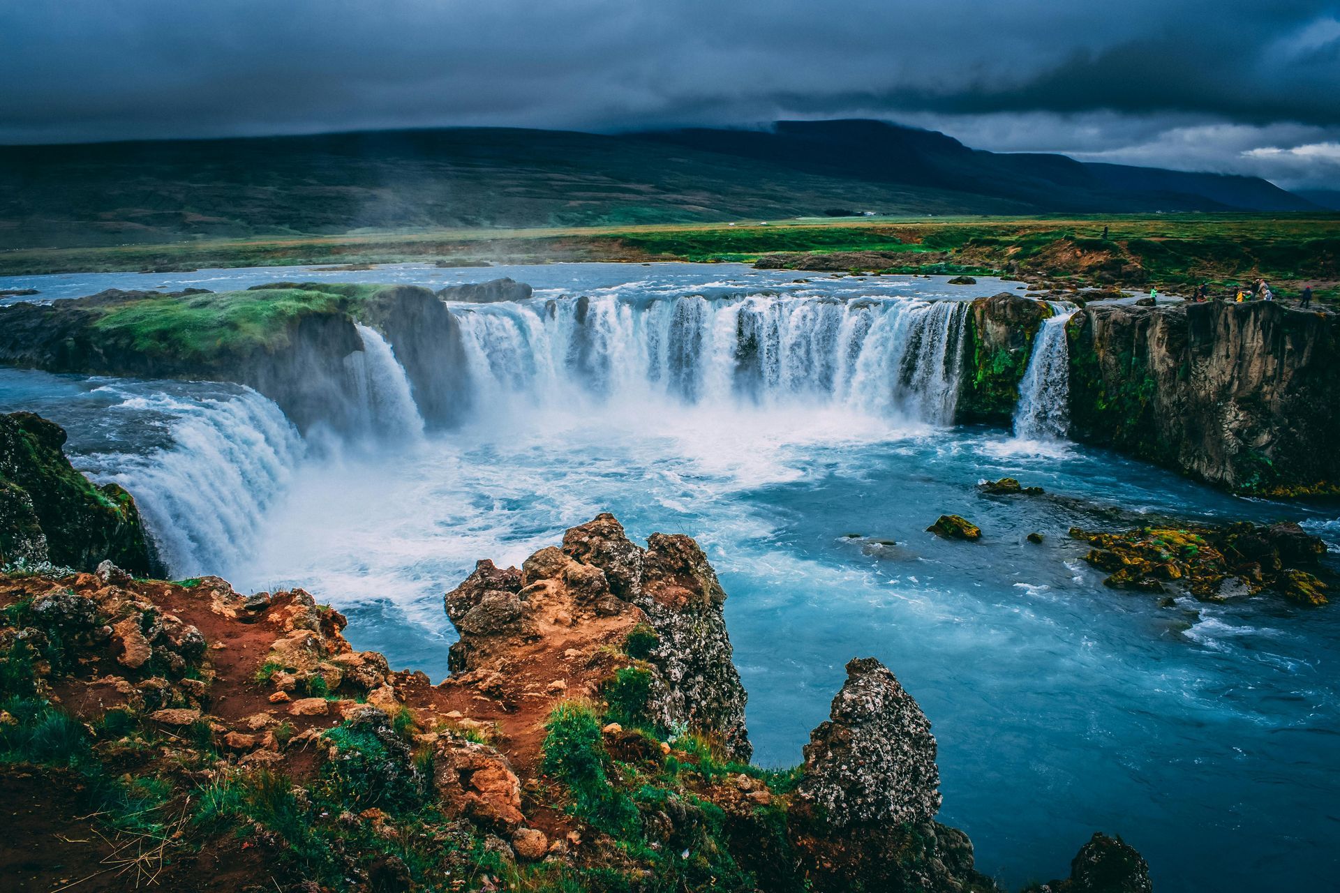 Waterfall cascading into turquoise water, with rugged, reddish-brown rocks and green vegetation, under a cloudy sky.