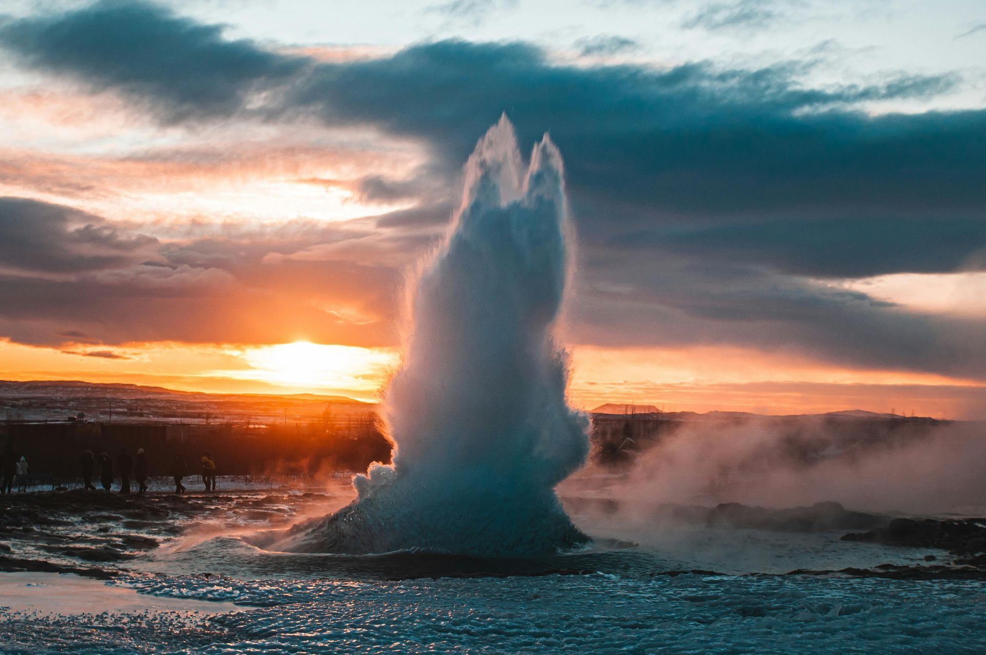 Geyser erupts, shooting water into the air against a sunset sky.