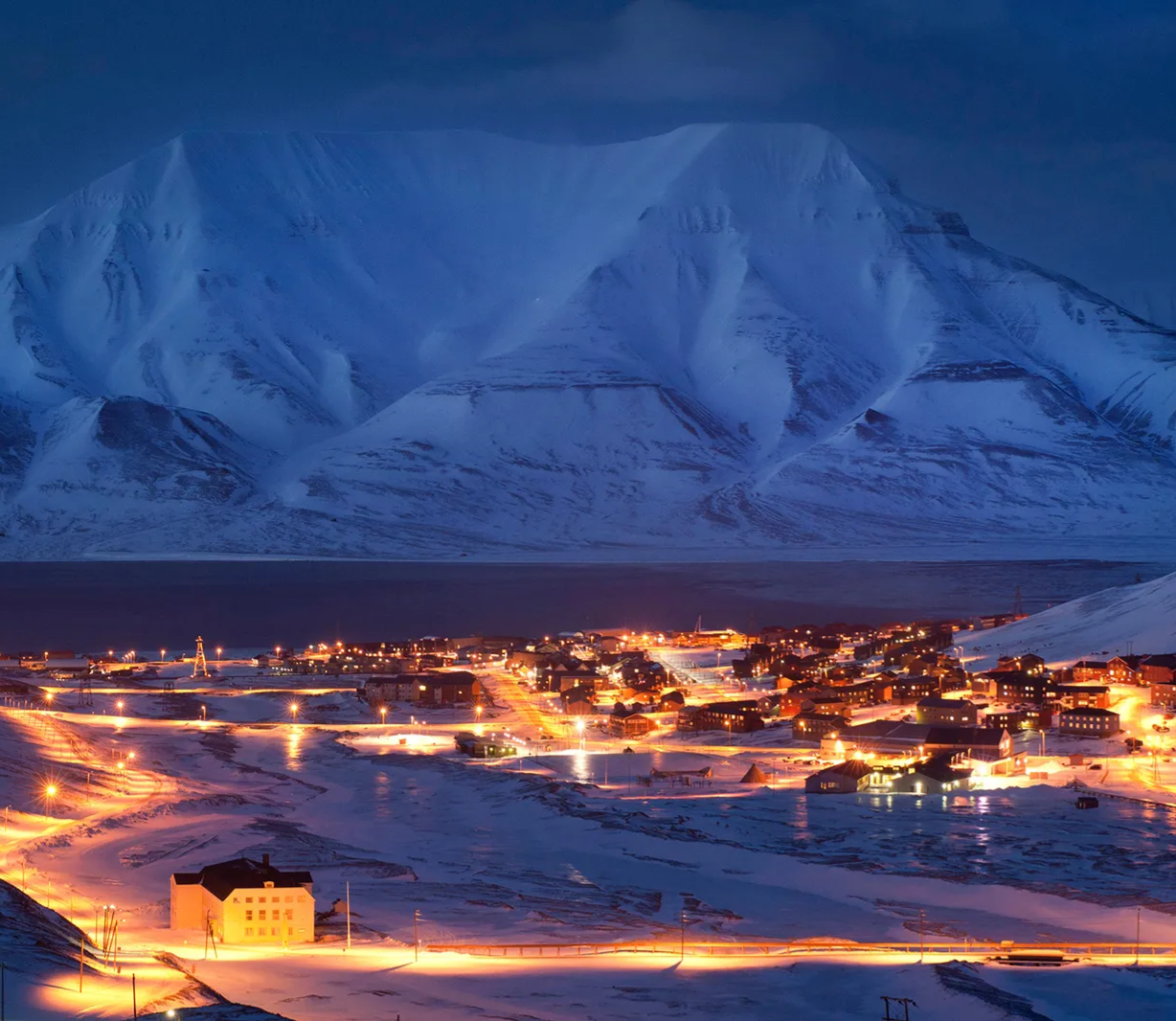 Aurora borealis in green and purple hues over snow-capped mountains and a dark body of water.