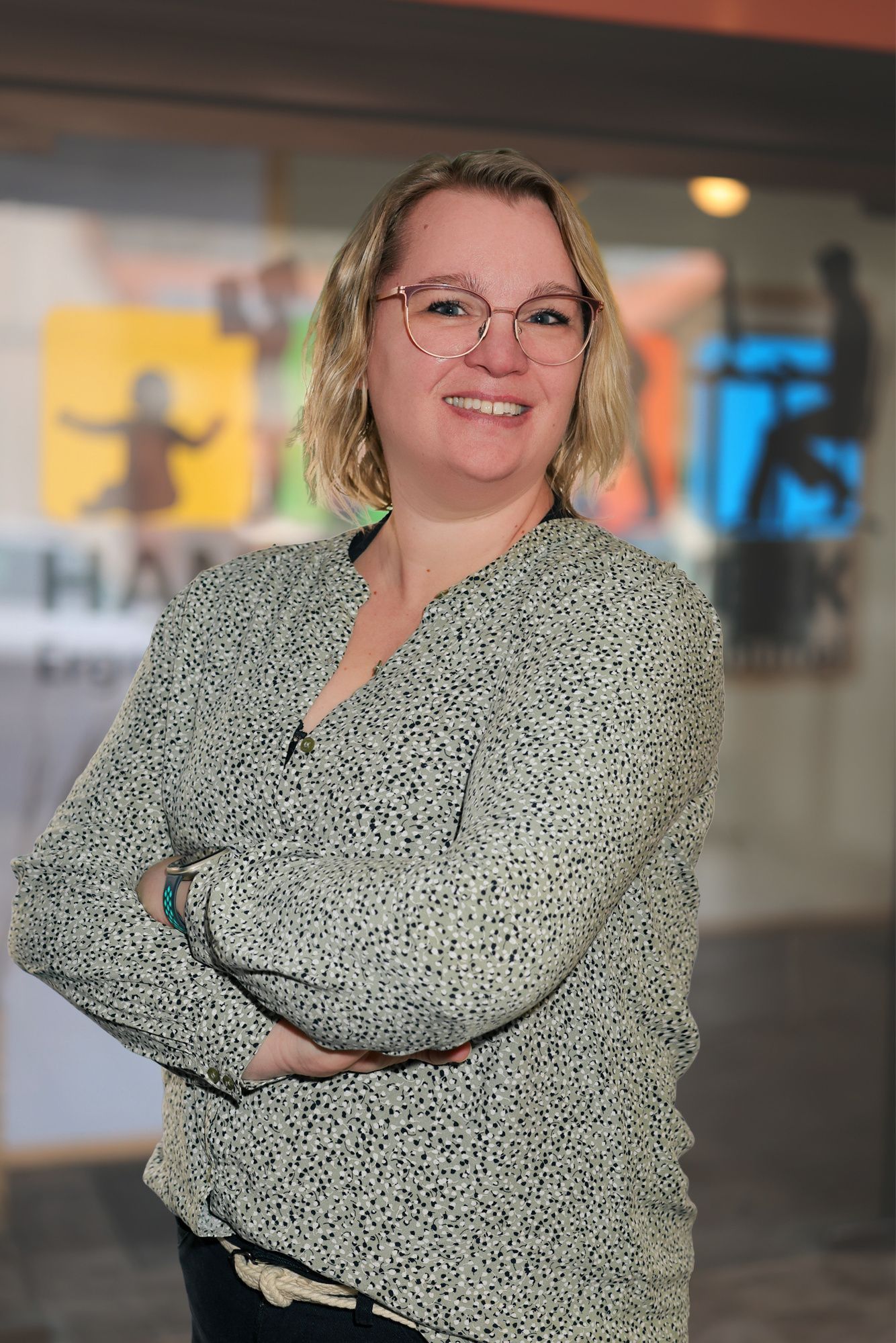 Frau mit Brille lächelt in einem Flur; trägt ein dunkles Oberteil mit goldenen Mustern.