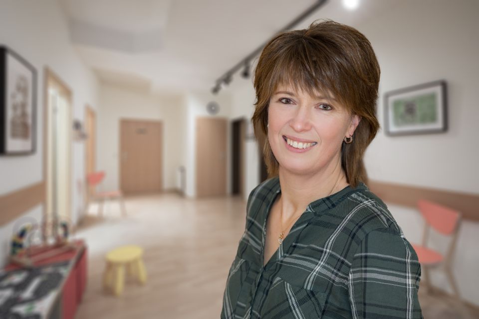 Frau im grün karierten Hemd lächelt in einem Flur mit Kunst und Stühlen.