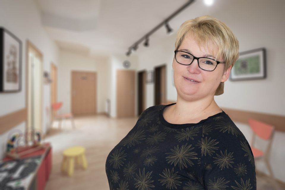 Frau mit Brille lächelt in einem Flur; trägt ein dunkles Oberteil mit goldenen Mustern.