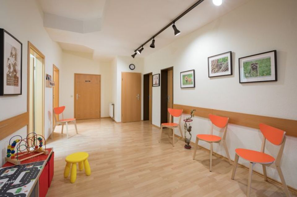 Wartezimmer mit orangefarbenen Stühlen, gerahmter Kunst und einem Kinderspielbereich.