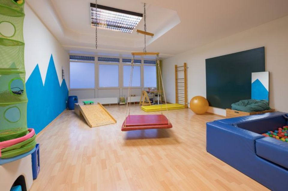 Therapieraum: blaues Bällebad, Holzrampe, Schaukel, Klettergerüste, an die Wand gemalte Berge.