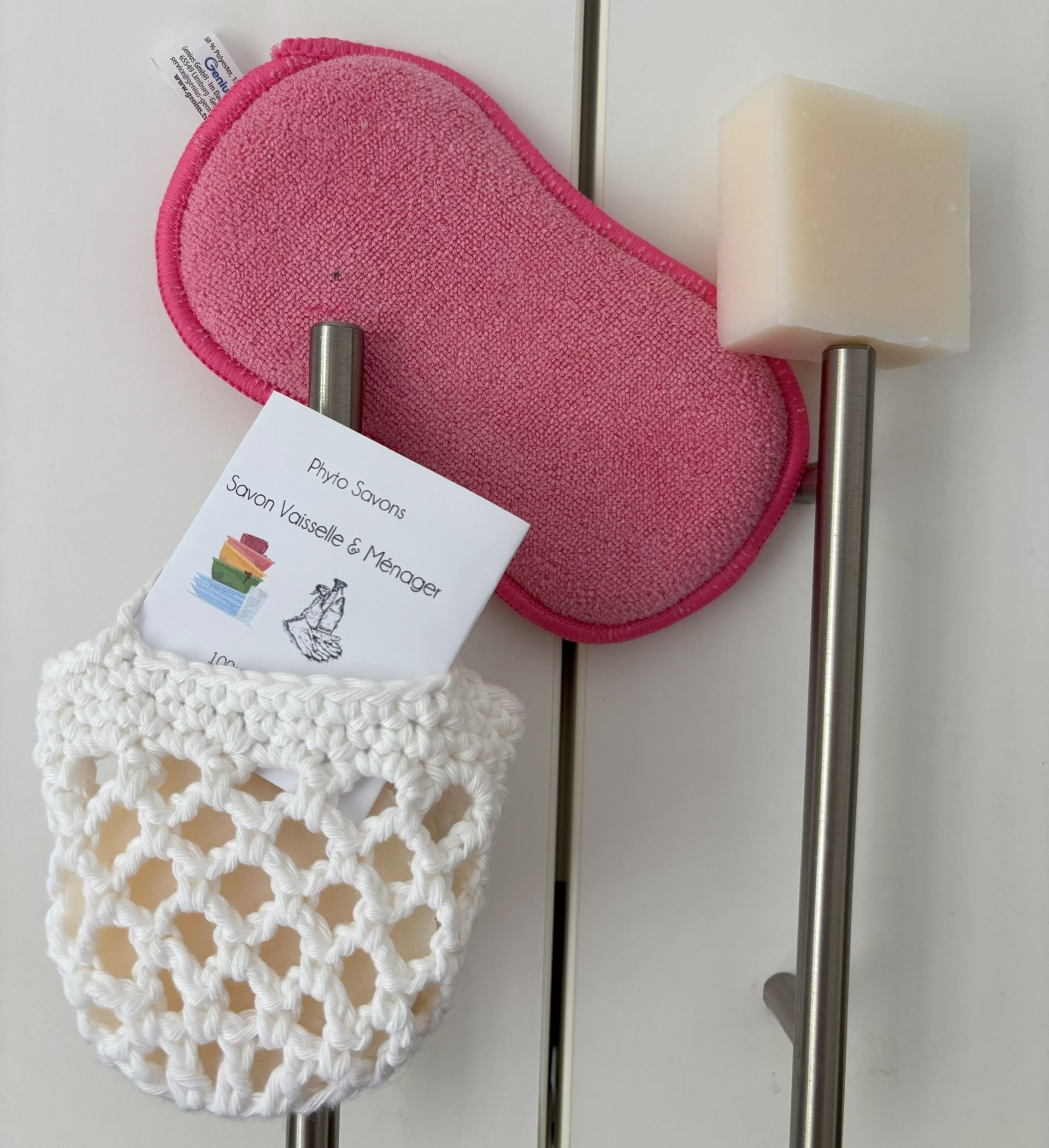 Porte-savon blanc au crochet, éponge rose et barre de savon suspendue à des tiges métalliques sur une armoire blanche.