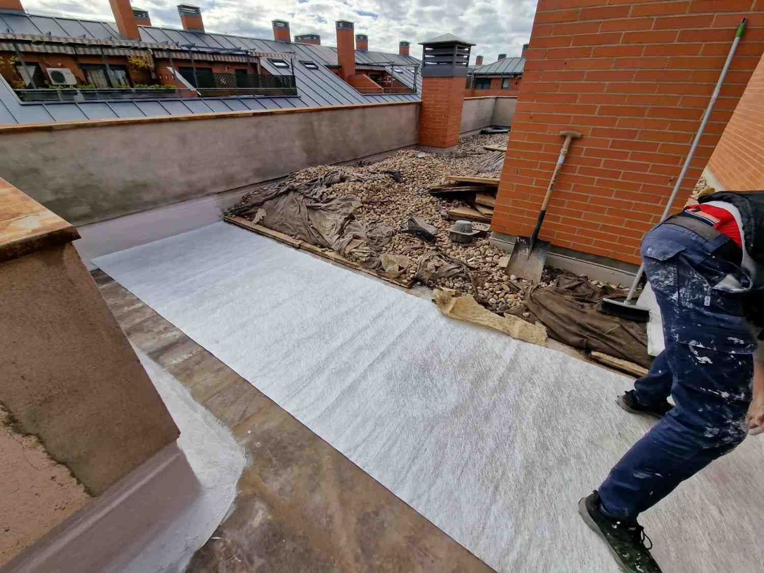 Trabajador instalando una membrana impermeabilizante blanca en una azotea plana junto a unas chimeneas de ladrillo