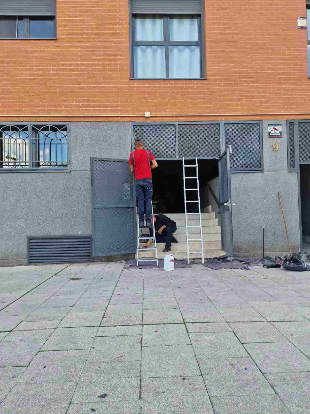 Dos trabajadores en la entrada de un edificio; uno subido a una escalera y otro de pie debajo con un cubo.