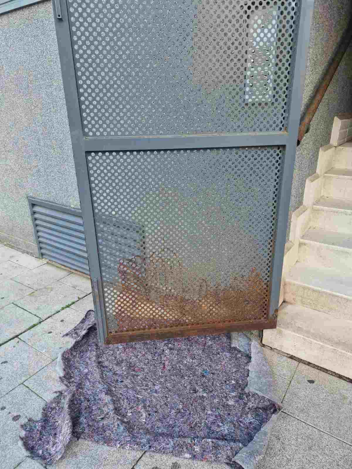 Una verja metálica rota con un gran agujero en el suelo, junto a unos escalones alicatados y la acera