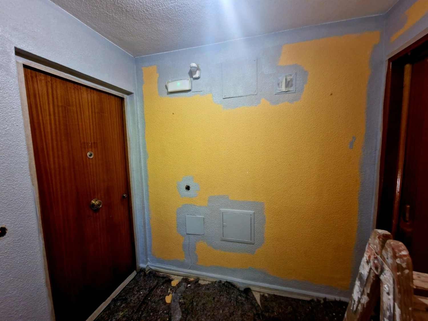 Pasillo pequeño con una pared amarilla, cajas de servicios, dos puertas de madera y algunos escombros de obra en el suelo.
