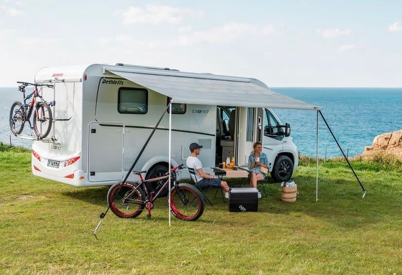 Una pareja está sentada bajo un toldo al lado de una autocaravana.