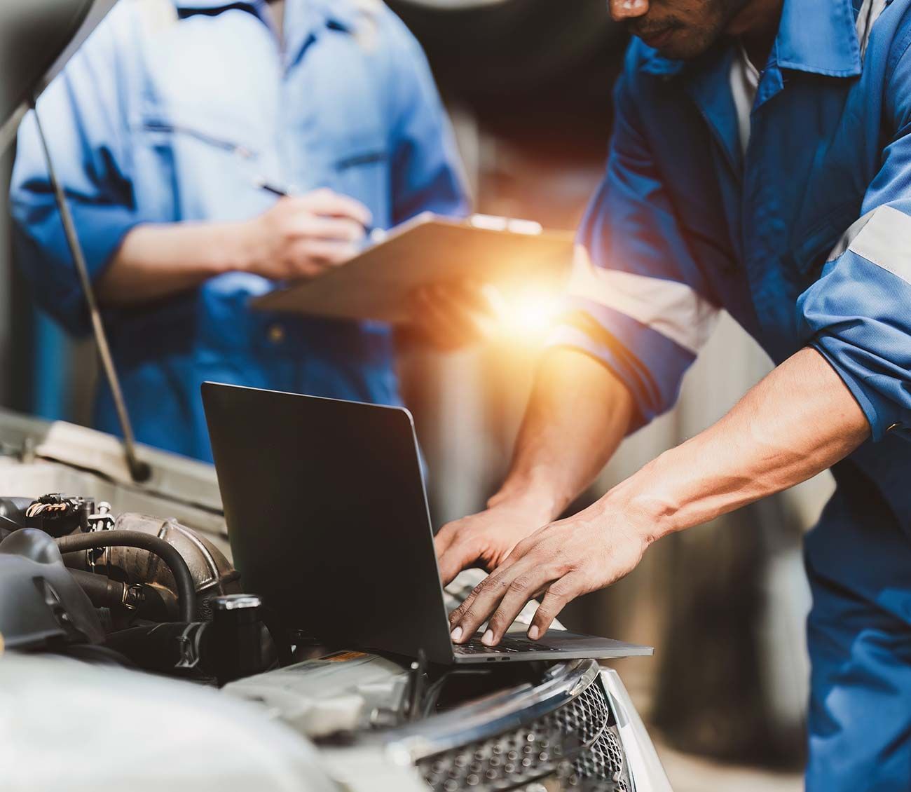 Mecánicos trabajando en una computadora portátil junto al motor de un automóvil en un garaje, con el resplandor de una linterna brillante.