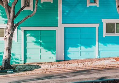 Maison turquoise avec portes de garage, un arbre et un trottoir devant.