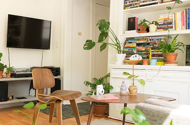 Salon confortable avec télévision murale, fauteuil en bois, table basse et plantes près d'une cheminée blanche.