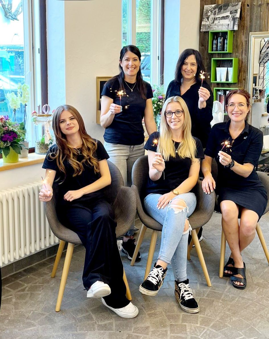 Fünf Frauen in schwarzen Outfits posieren und lächeln in einem hellen Salon, einige sitzen, andere stehen mit Getränken.