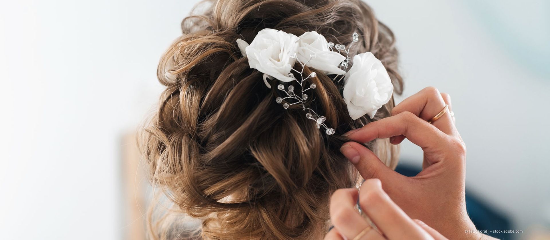 Eine Stylistin arrangiert eine hochgesteckte Frisur für braunhaarige Frauen mit weißen Blumen und Perlenhaarnadeln.