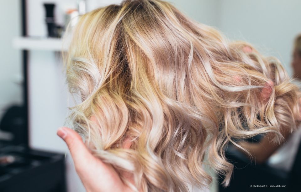 Eine Hand hält schulterlanges, blondes, gewelltes Haar mit weichen Locken und Highlights.