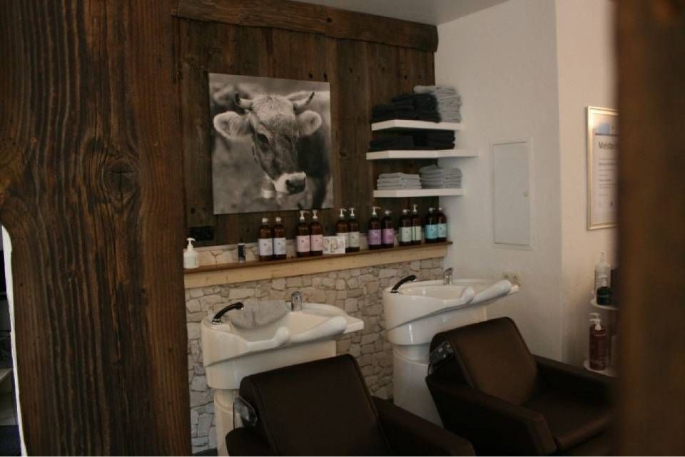 Ein Friseursalon-Waschplatz mit zwei Waschbecken, dunkelbraunen Stühlen, einem Kuhporträt an einer Holzwand und weißen Ablageregalen.