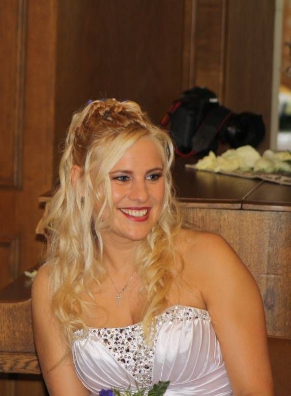 Eine lächelnde Frau in einem trägerlosen weißen Brautkleid mit paillettenbesetztem Oberteil, die ihr blondes Haar halb hochgesteckt trägt.