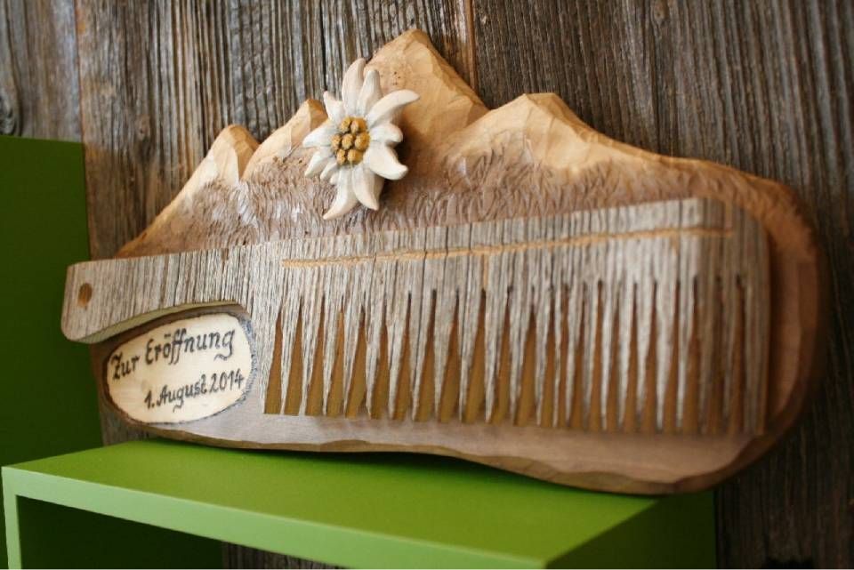 Eine hölzerne Kammschnitzerei, montiert auf einer bergförmigen Plakette, mit einer Edelweißblume und der Datumsangabe „1. August 2014“.