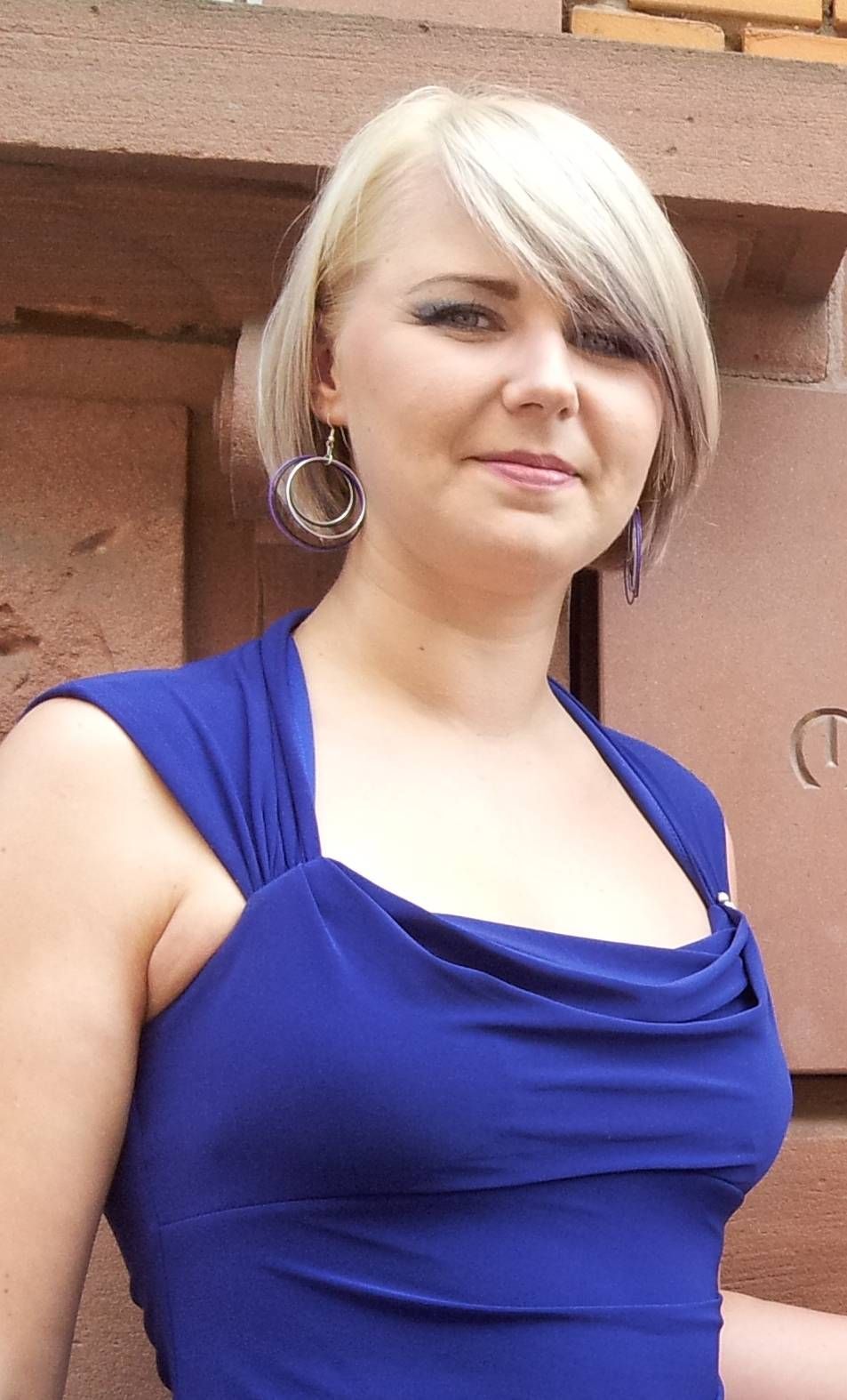Eine Person mit kurzen blonden Haaren und silbernen Creolen trägt ein hellblaues, ärmelloses Kleid vor einer Backsteinmauer.