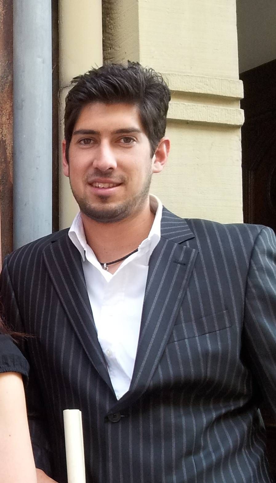 Eine Person mit dunklem Haar und hellem Bart trägt ein Nadelstreifen-Sakko und ein weißes Hemd mit Kragen und steht an einer Wand.