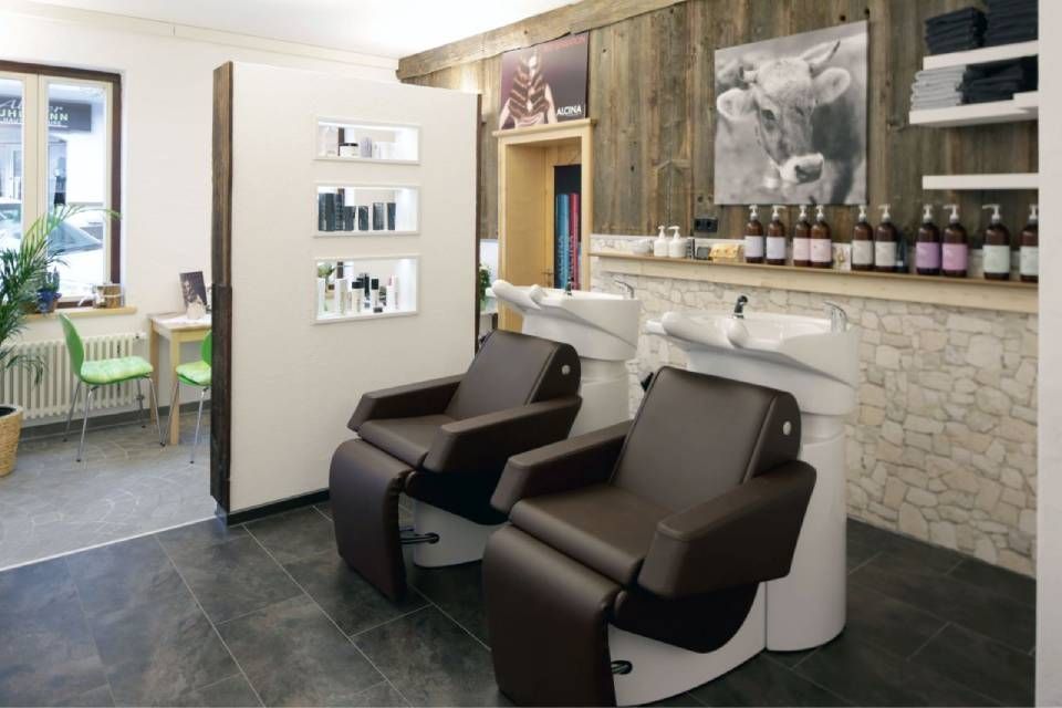 Ein Friseursalon ist mit zwei braunen Shampoo-Stühlen, einer weißen Trennwand mit Ausstellungsregalen und einer rustikalen Holzwand mit Kunstwerken ausgestattet.