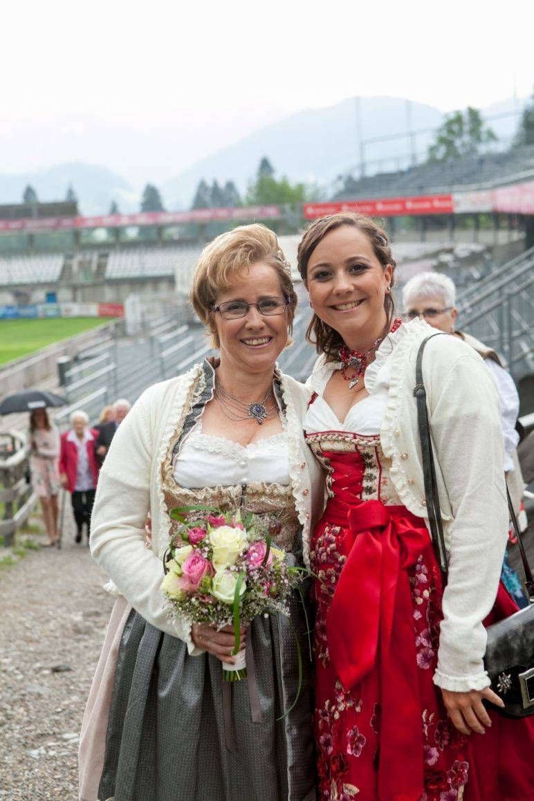 Zwei Frauen in traditionellen Dirndlkleidern mit weißen Strickjacken posieren für ein Foto in einem Freiluftstadion.