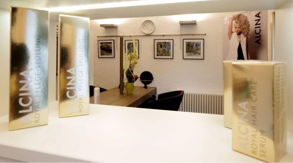 Alcina-Haarpflegeprodukte in goldener Verpackung stehen auf einem Tresen vor einem Spiegel und einem Tisch in einem Salon.