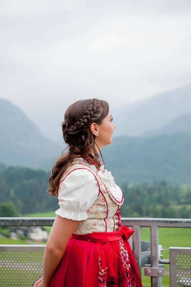Eine Person in einem rot-weißen Dirndl steht auf einem Balkon mit Blick auf eine Berglandschaft unter bewölktem Himmel.