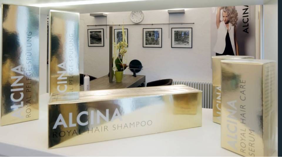 Goldfarbene Haarpflegeprodukte der Marke Alcina, darunter Shampoo und Serum, werden in einem hellen Friseursalon auf einem weißen Regal präsentiert.