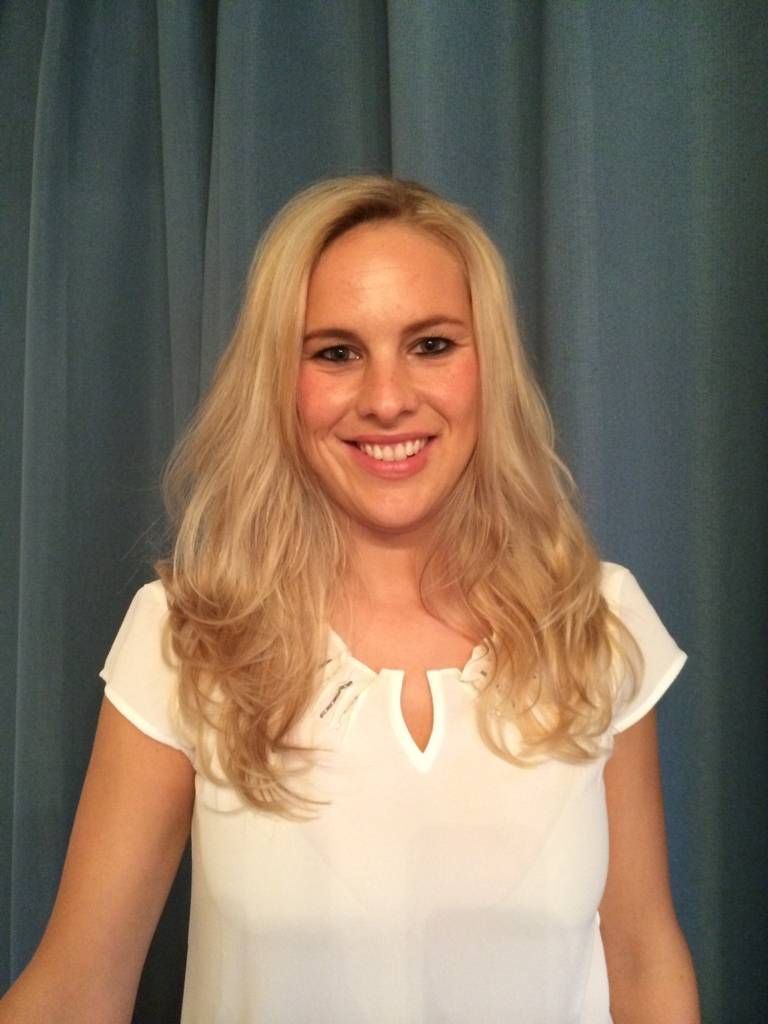 Lächelnde Person mit langen blonden Haaren und einem weißen Kurzarmtop vor einem blauen Vorhang.