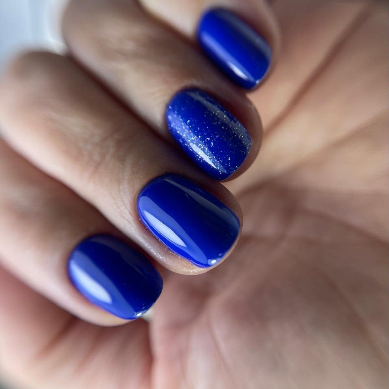 Cuatro dedos con uñas cortas y redondeadas pintadas de un azul real vibrante, y la segunda uña con un sutil brillo plateado.