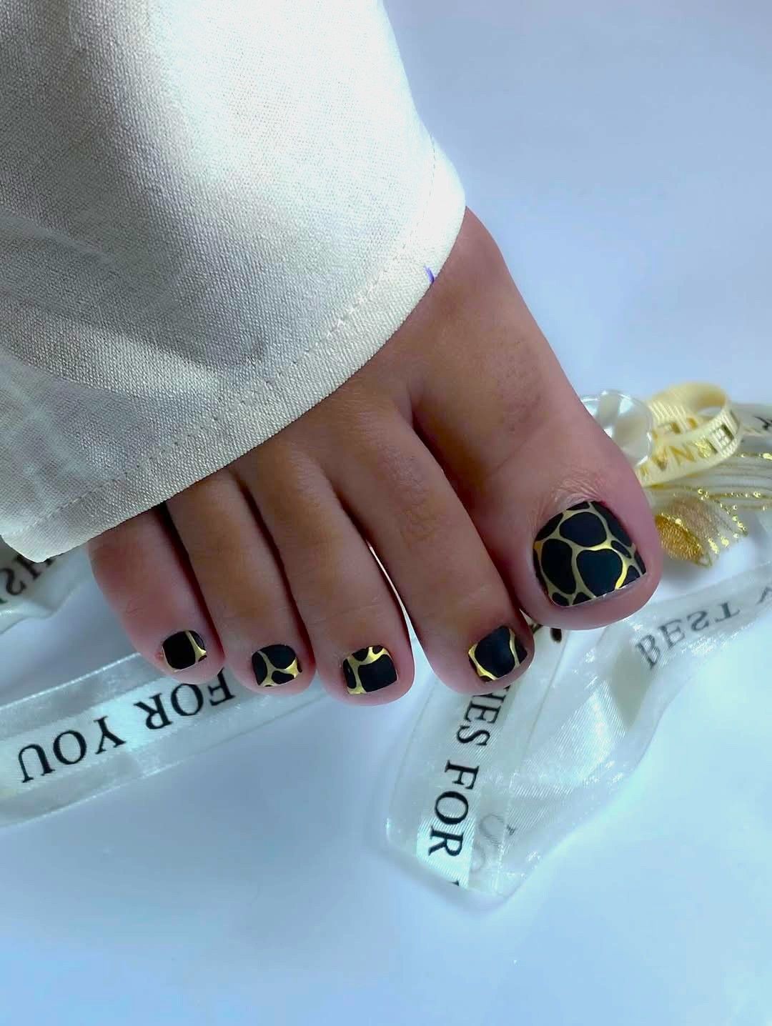 El pie de una persona con las uñas pintadas con un estampado de jirafa en negro y dorado, sobre un fondo blanco.