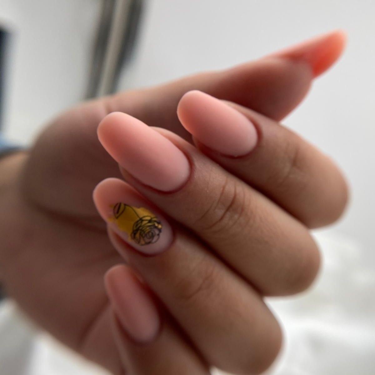 Mano con una manicura de color melocotón suave, con uñas de forma ovalada y un pequeño detalle de rosa en una de ellas.