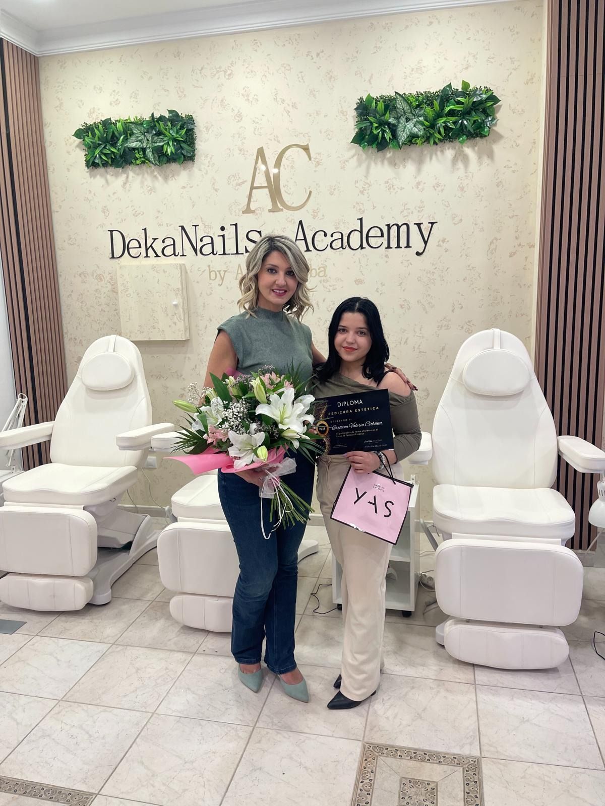 Dos personas están de pie juntas en una academia de manicura, sosteniendo flores y un pequeño cartel frente a sillones 