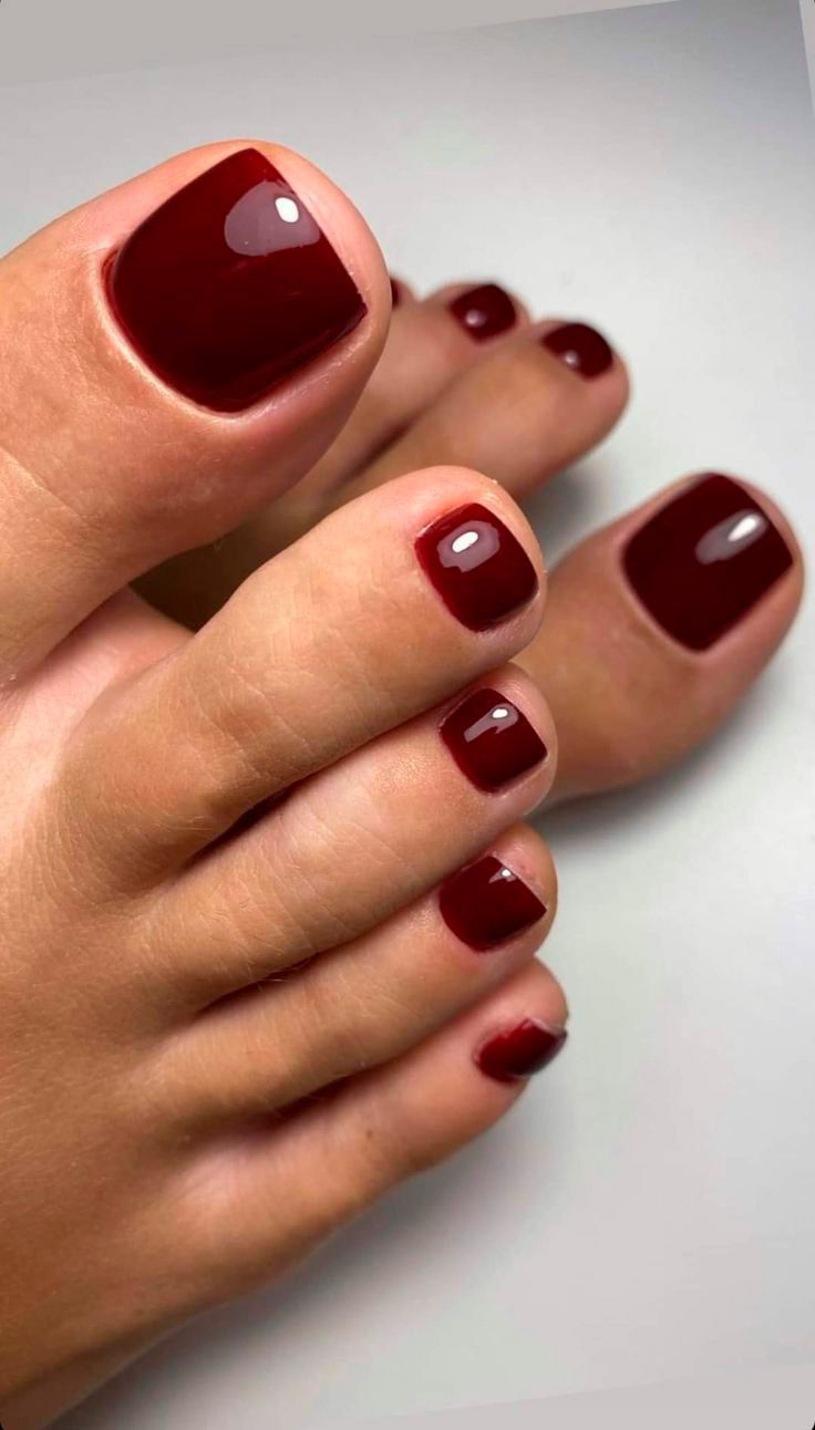 Primer plano de los dedos de los pies con esmalte de uñas rojo oscuro brillante sobre una superficie blanca.