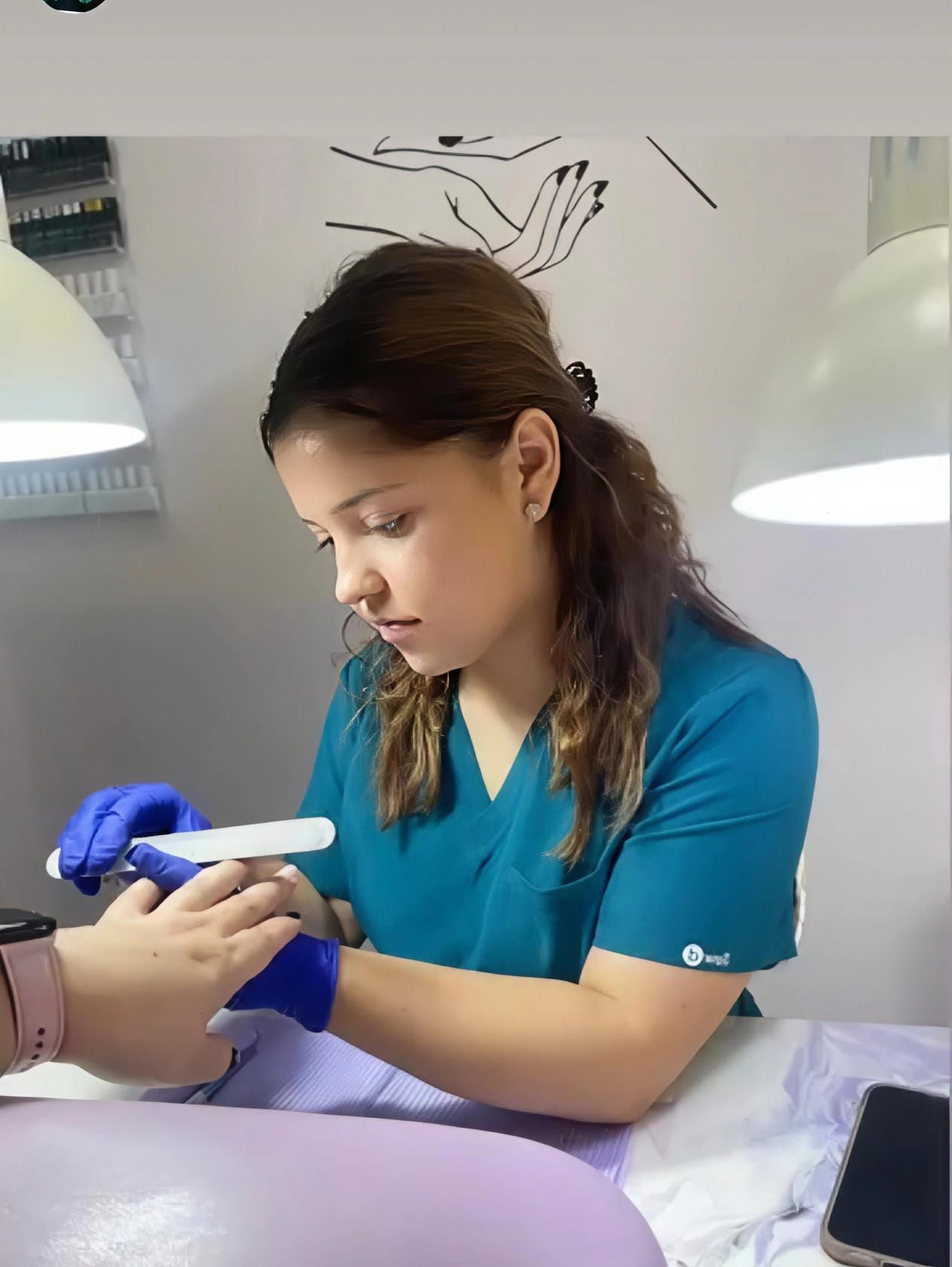Una persona vestida con bata y guantes azules le lima las uñas a una clienta en un salón de manicura muy iluminado.