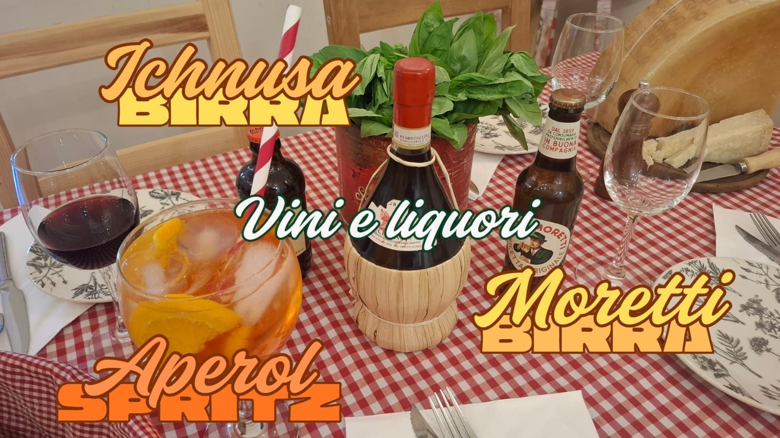 Mesa con bebidas: Aperol Spritz, vino tinto, cervezas Ichnusa y Moretti y una botella de vino cubierta con paja; mantel a cuadros rojos y blancos.