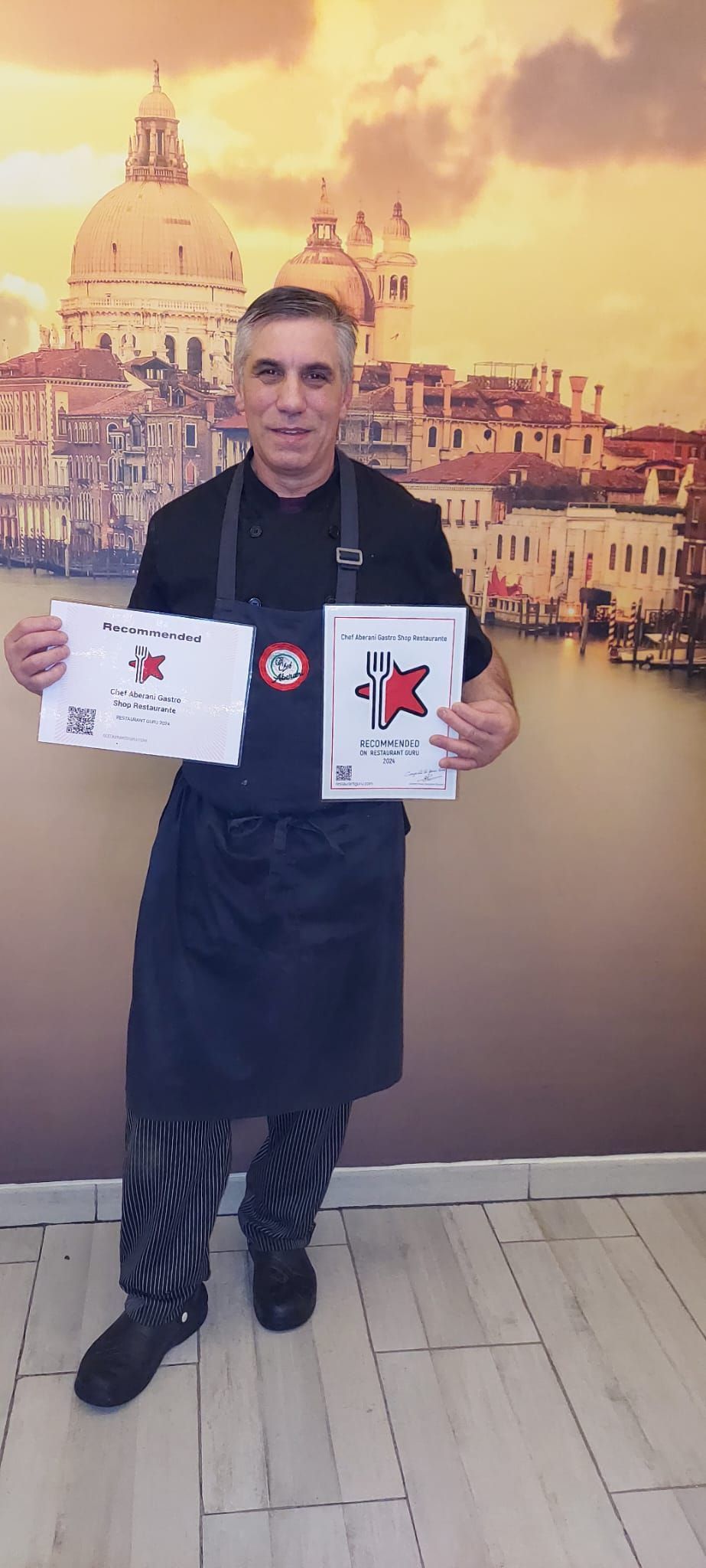 Un hombre con delantal sostiene dos papeles delante de una imagen de Venecia.