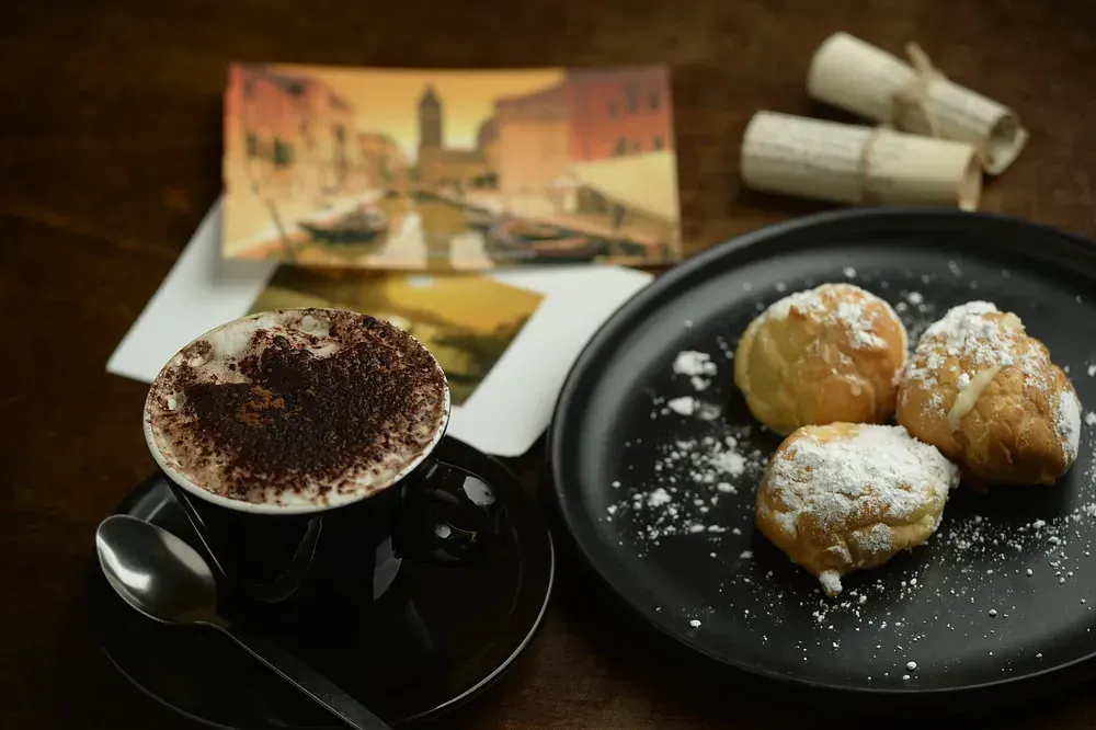 Una taza de capuchino junto a un plato de pasteles con azúcar glas