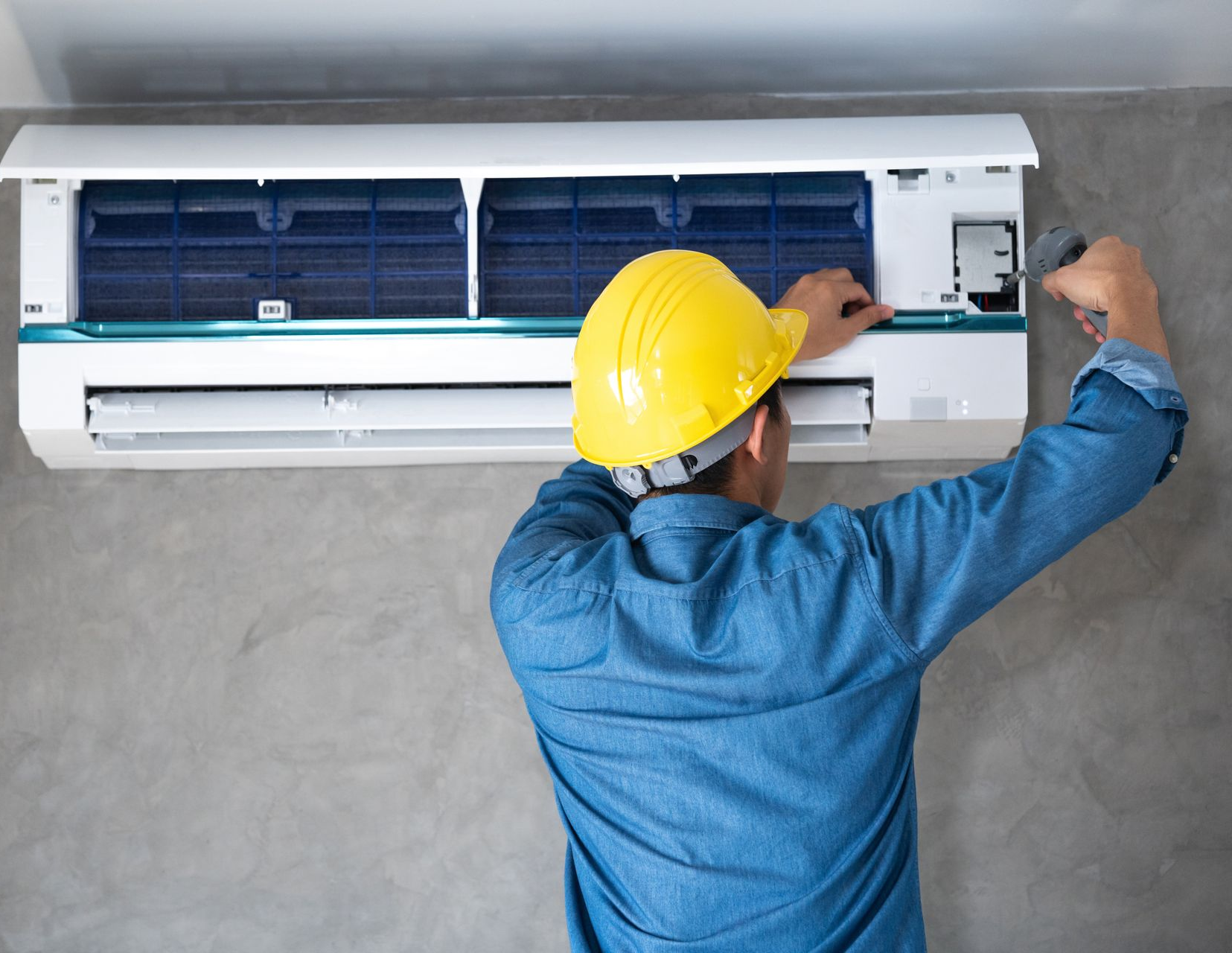 Un technicien portant un casque de chantier répare un climatiseur mural à l'intérieur.