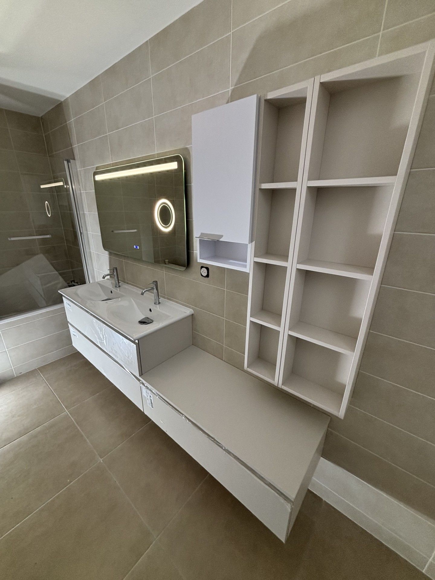 Salle de bains moderne avec lavabo suspendu, miroir éclairé et étagères de rangement dans une pièce carrelée.