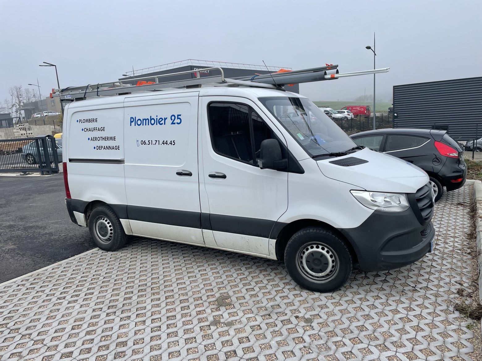 Fourgonnette de service avec échelle sur le toit, garée sur un parking goudronné à côté d'une voiture.